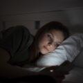 Uma mulher jovem usando o celular na cama, ou seja, precisando de um detox digital antes de dormir.